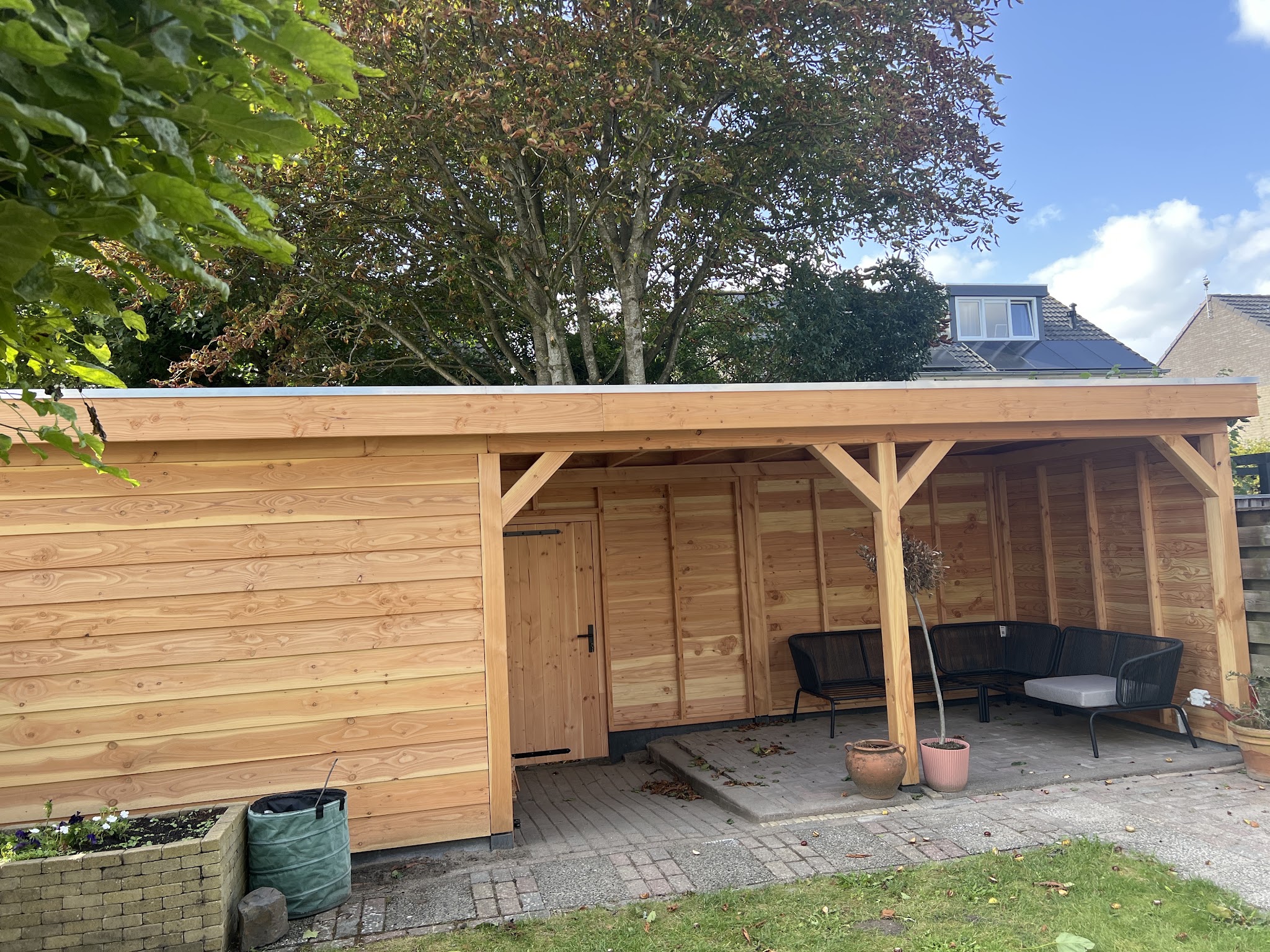 Houten schuur met overkapping en loungeset in een tuin met bomen